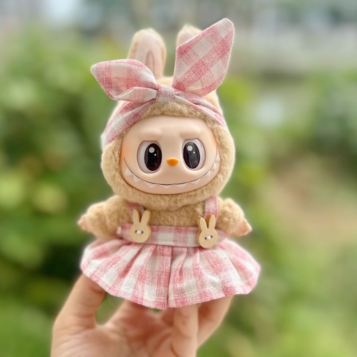 Cute Labubu plush dressed in Zibuya’s pink gingham dress set, featuring bunny-shaped buttons and a matching bow headband, held outdoors with greenery in the background.