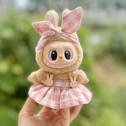 Cute Labubu plush dressed in Zibuya’s pink gingham dress set, featuring bunny-shaped buttons and a matching bow headband, held outdoors with greenery in the background.